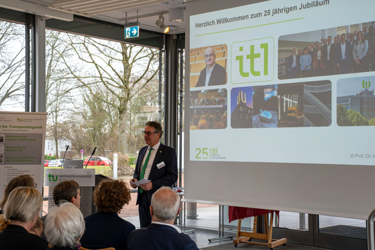 Professor Uwe Clausen neben einer Folienpräsentation zur Begrüßung der Veranstaltung, während die Teilnehmer ihm aufmerksam zuhören.