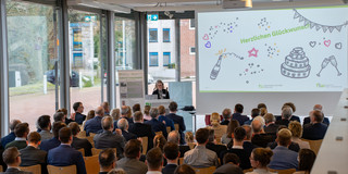 Presentation by Professor Kirchheim, standing in the middle with a microphone and a slide presentation in the background.