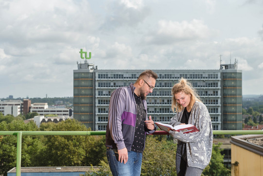 A male and a female student are looking into a book together, in the background the Math Tower can be seen in its full length.