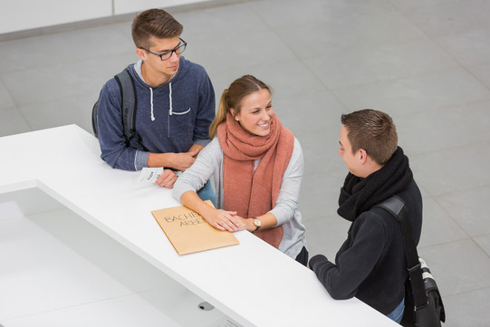 Studierendensekretariat Drei Personen stehen neben einenander an einer Theke. Die Person in der Mitte lacht die Person rechts an und hat ihre Hände auf einen großen Umschlag liegen. Die Person links hat einen kleinen Zettel mit einer Wartenummer in der Hand.