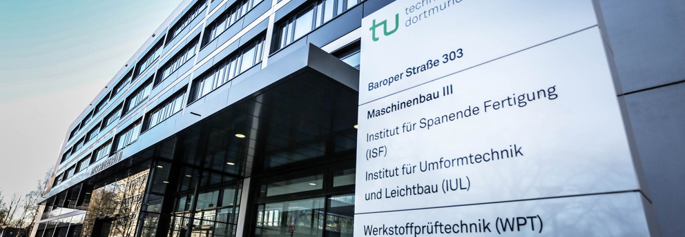 Maschinenbau-Gebäude III: Ein modernes grau-schwarzes Gebäude aus Glas mit Vordach, seitlich von unten fotografiert. Rechts vor dem Gebäude ein Schild mit der Aufschrift "Technische Universität Dortmund, Baroper Straße 303, Maschinenbau 3" und weiteren Instituten und Einrichtungen.