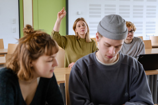 Students in the seminar room are sitting at the tables. One student give a hand signal.