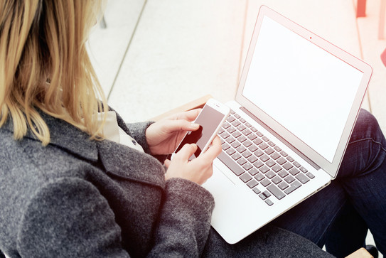 Frau mit Laptop auf dem Schoß, hält ein Smartphone in den Händen.