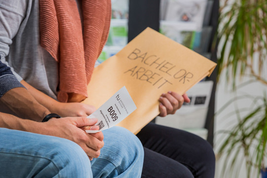 Zwei Personen sitzen nebeneinander. Man sieht einen Ausschnitt von Knie bis Brust. Die hintere Person hat einen großen Umschlag mit der Aufschrift „Bachelor-Arbeit“ in der Hand, die vordere Person hat einen kleinen weißen Zettel mit einer Wartenummer, einem Code und dem TU-Logo in der Hand.