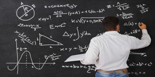 A man is seen from behind. He is writing formulas on a chalkboard