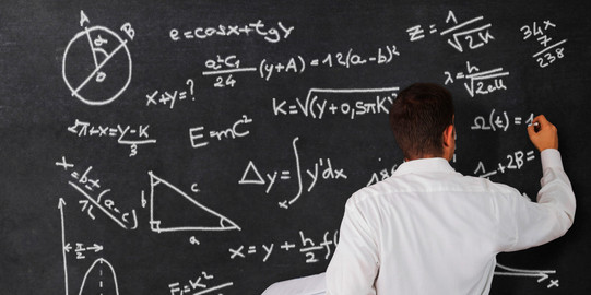 A man is seen from behind. He is writing formulas on a chalkboard