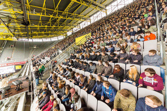 Studierende des ersten Semesters sitzen bei der Erstsemesterbegrüßung auf den Rängen im Stadium und schauen seitlich einem Showact zu.